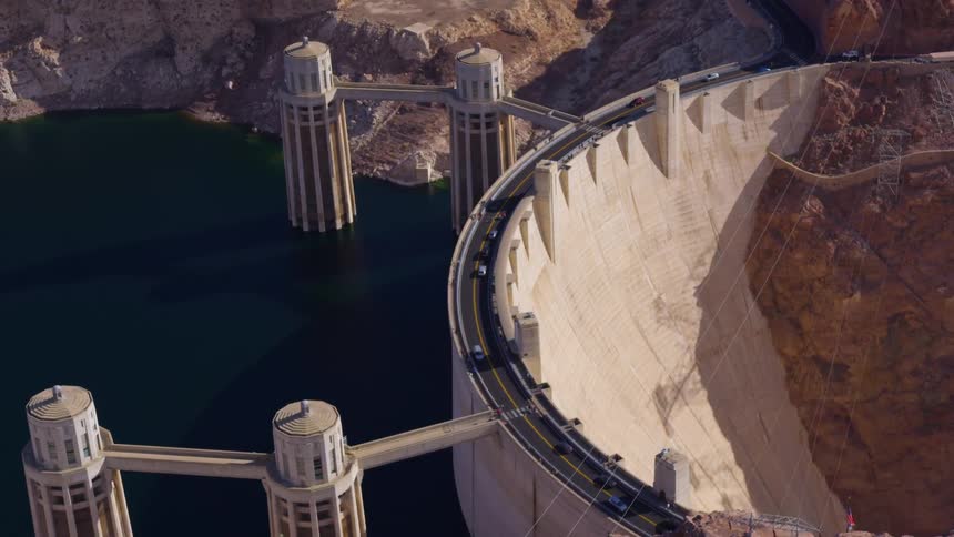 An aerial shot shows a wide curved concrete dam with a road running across its top on one side of the dam cars move along the road while a large serene body of water rests on the other