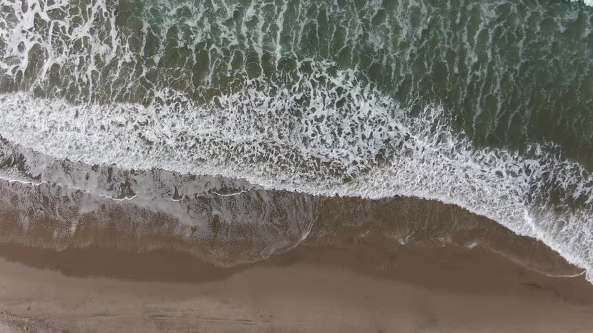 The video shows a highangle shot of ocean waves rolling into a sandy beach the foamy white surf of each wave spreads out across the brown sand before receding back into the deeper dark green water