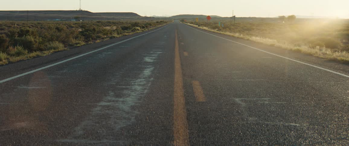 The suns golden rays illuminate a long straight desert highway highlighting wet patches on the asphalt and distant hills