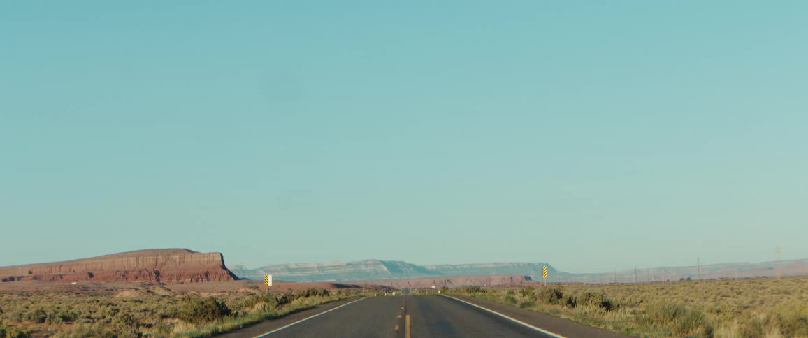A shot from a moving car shows a long empty desert highway stretching towards distant mesas under a clear blue sky
