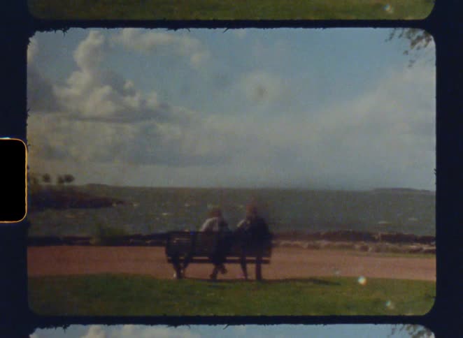 Three people sit on a bench facing the ocean in vintage film style