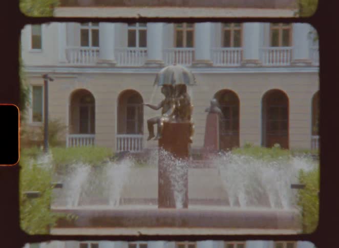Tourist outside walking near a building with a fountain that has a statue in the middle water sprinkling at midday and grasses surrounding the building
