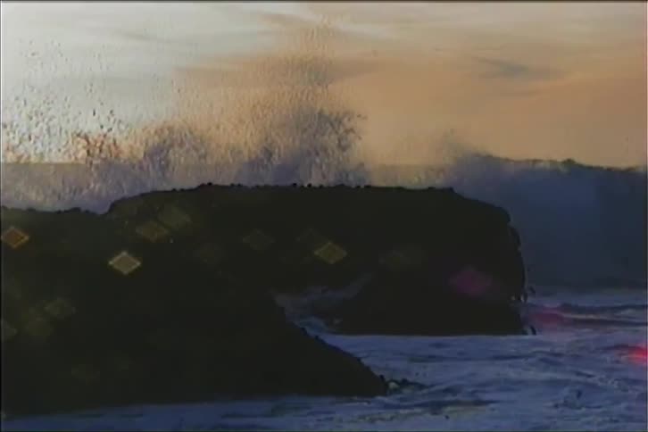 This grainy video shows powerful waves crashing against a rocky shoreline during a sunset the camera zooms in capturing the foam and spray of the surging water as it surrounds the dark rocks