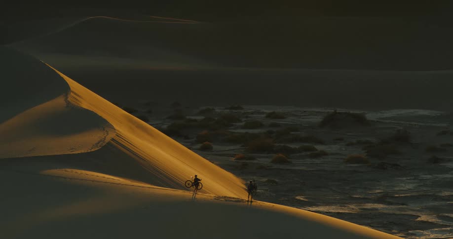 This wide cinematic shot shows two people walking on the edge of a large sand dune at sunset the low sun casts a beautiful golden light on the dune creating a stark contrast with the darker landscape in the background