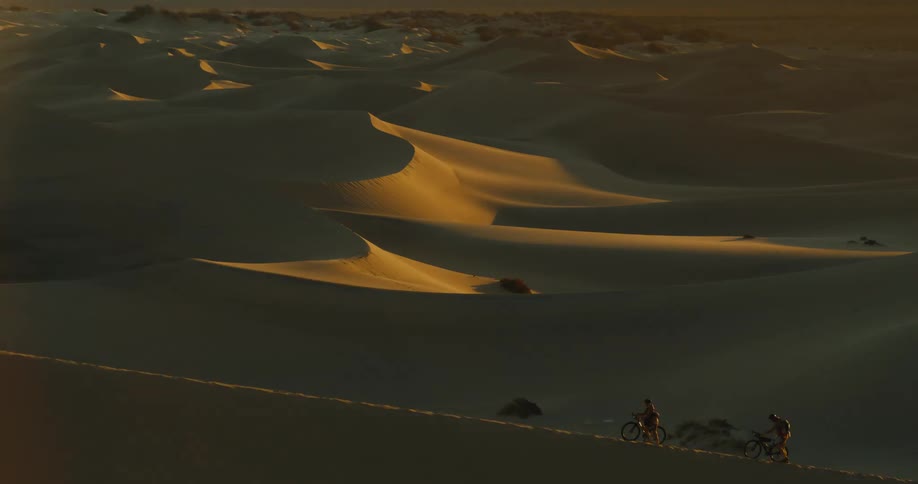 Two people walking with their bicycles across sand dunes in a desert at sunset
