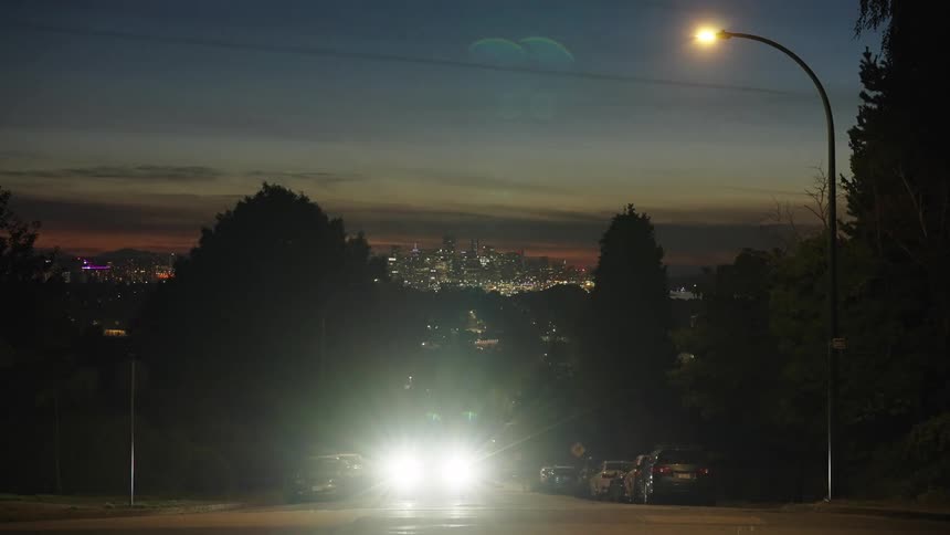 This timelapse video shows a suburban street at dusk with cars headlights creating bright streaks of light as they pass in the background the city skyline is visible slowly becoming more illuminated as night falls