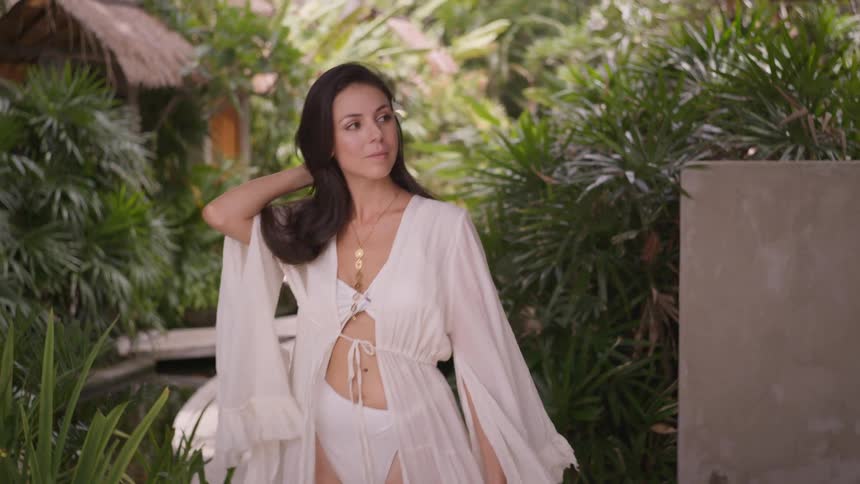 Woman wearing a white beach coverup walks among tropical vegetation with a flower in her hair