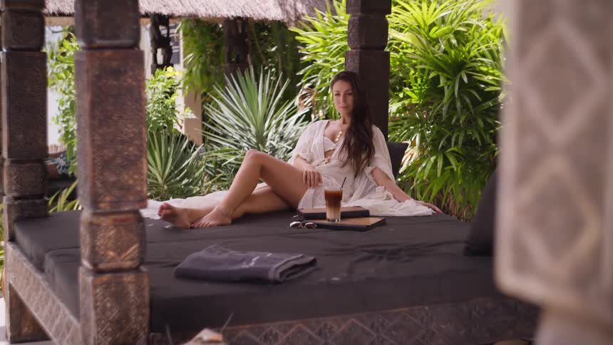 The video begins with a wide static shot of a young woman sitting on a daybed in a tropical setting with lush greenery in the background the camera then slowly zooms in on her as she reaches for and takes a drink from a glass on the table next to her
