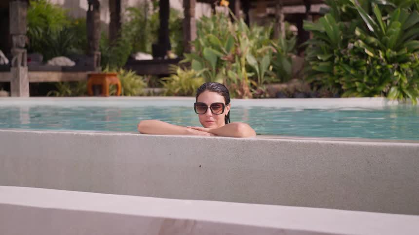 A woman swimming in a pool at midday resting by the edge as she lifts her glasses from her face