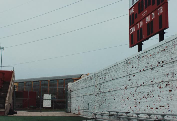 Red stadium scoreboard appears above a worn white wall with metal bleachers and a yellow train passing in the background
