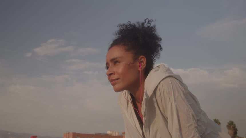 Close up of a young black woman listening to music with wired earphones while standing on a rooftop during daytime