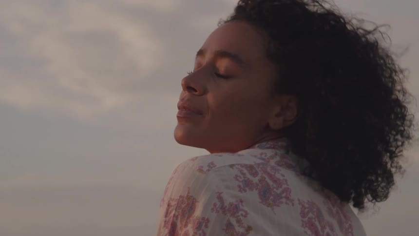 A young woman with curly hair smiles contentedly turning her face to feel the warm glow of the sun on a cloudy day