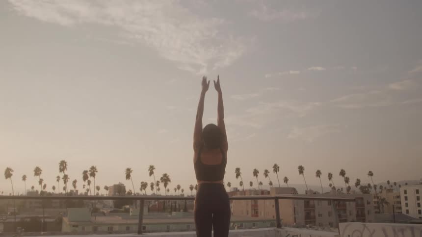 A person in athletic wear stands on a rooftop facing away from the camera they raise their arms above their head in a stretching or yoga pose silhouetted against the bright hazy sky