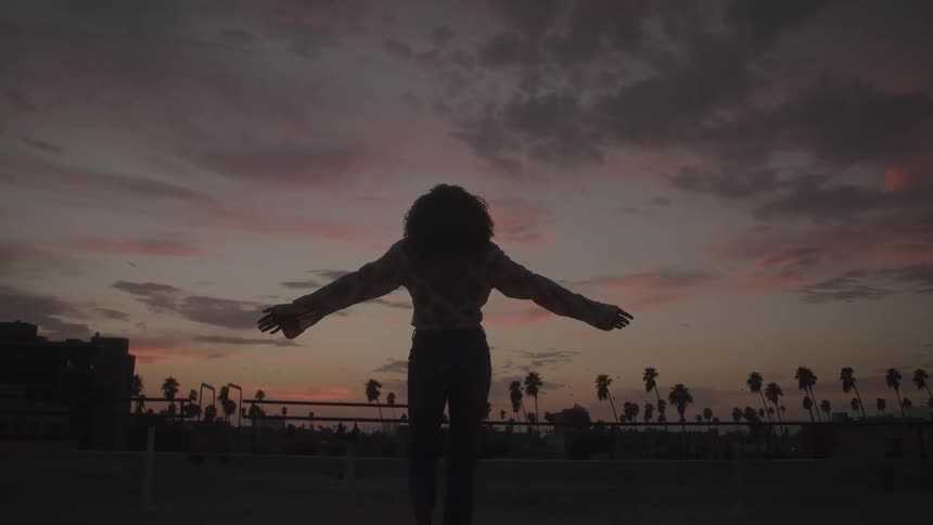 A young woman stands on a rooftop with buildings and palm trees in the distance she puts her arms out and looks at the sky as the sun sets