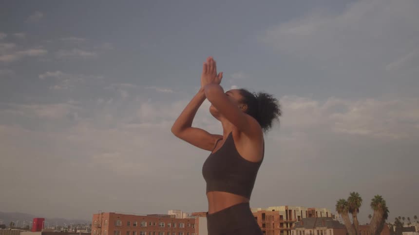 A woman practices yoga in a prayer pose on a rooftop at sunset