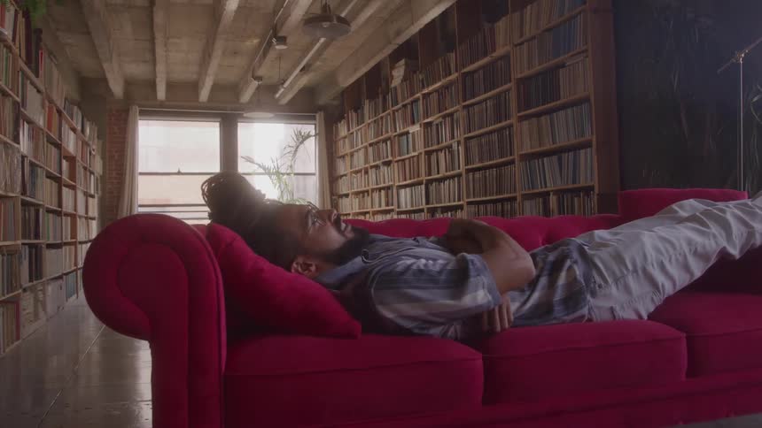 The man is lying on a red couch hes relaxing in a room filled with bookshelves