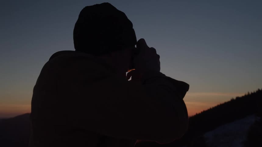 A man taking photographs of his surroundings with a camera as the orange setting sun glows in the sky