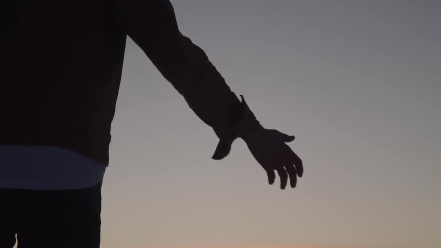A person is silhouetted against a setting sun as they face the right side of the screen they hold their arm out their hand open and palm up as if to catch the suns fading light