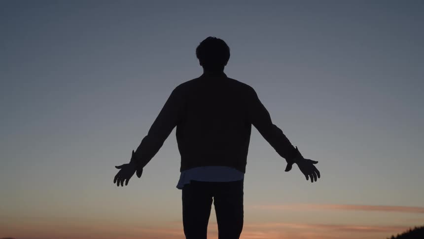 A silhouetted man stands on a mountain with his back to the camera with his hands down to his sides and then slowly raises his arms out wide with a sunset and a mountain range in the distance