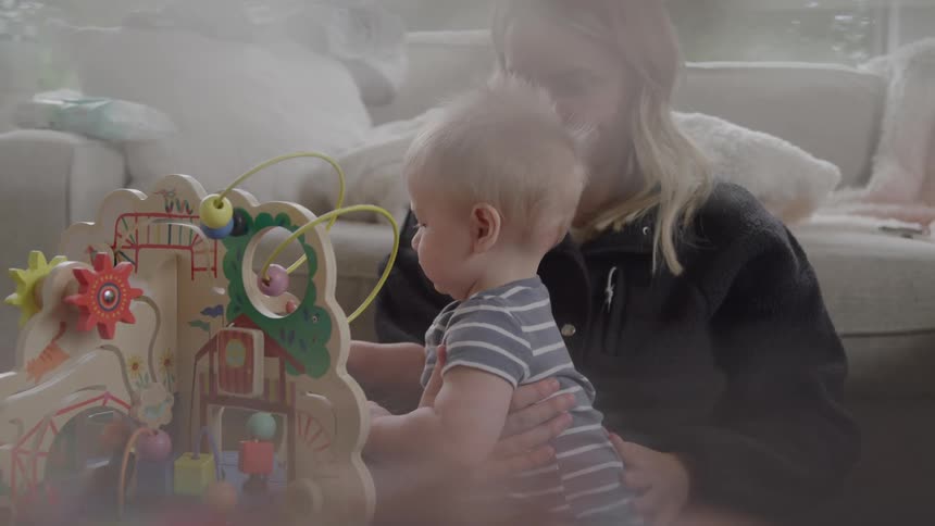 A young mother rubbing her babys back as he plays with toys on the floor in the living room during daytime