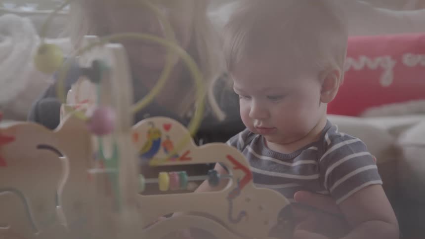 A baby sitting next to a female adult leans over a wooden activity toy brows knitted in concentrated curious exploration