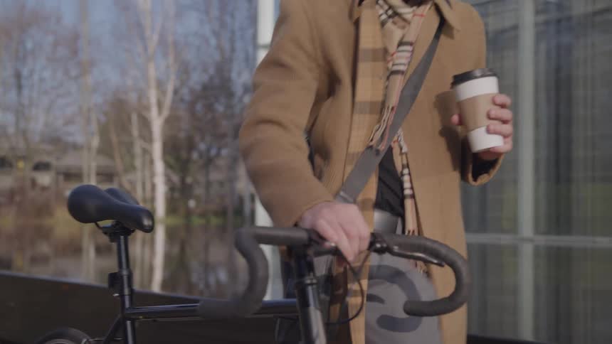 A person wearing a long camelcolored coat and a patterned scarf is holding a takeaway coffee cup while walking a black bicycle