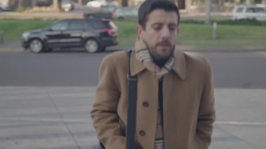 A man with dark hair and a beard wearing a brown coat and a striped scarf walks down a flight of stairs he carries a black messenger bag over his shoulder