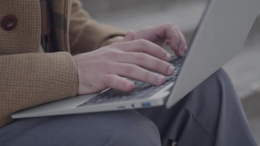 A persons hands are on a laptop and typing on the keyboard while the person is sitting on a stair