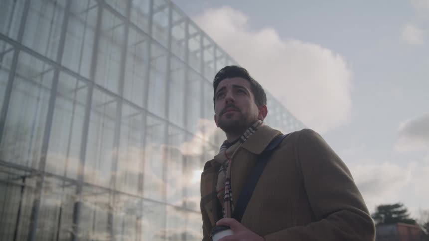 A low angle view shows a man with a beard wearing a camel coat and a patterned scarf looking up with a thoughtful expression while standing next to a modern glass office building