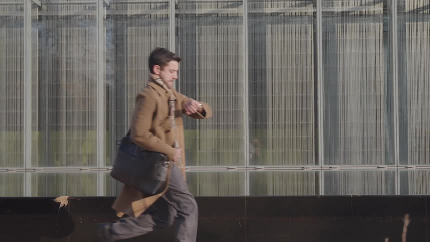 Man in a brown jacket running along the sidewalk on a sunny day carrying a handbag