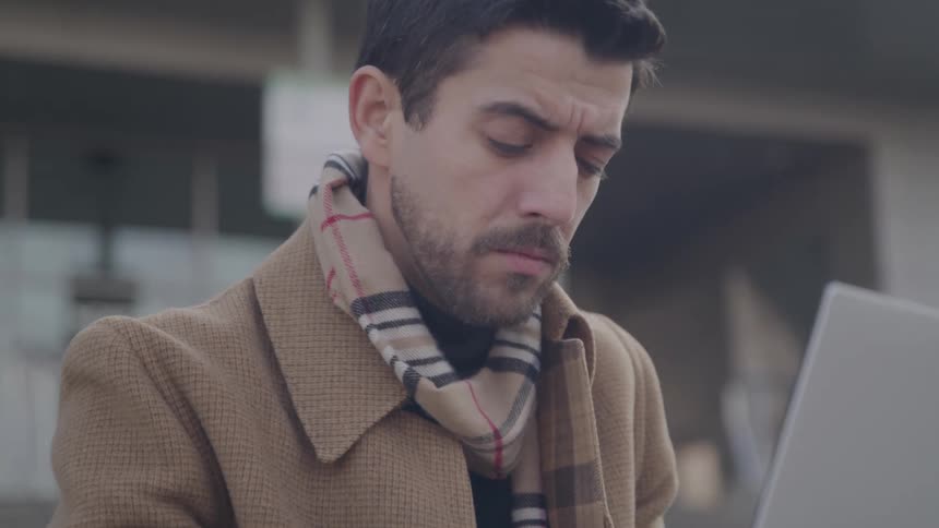 A man with a beard wearing a brown coat and a patterned scarf looks down at a laptop screen with a serious and concentrated expression on his face