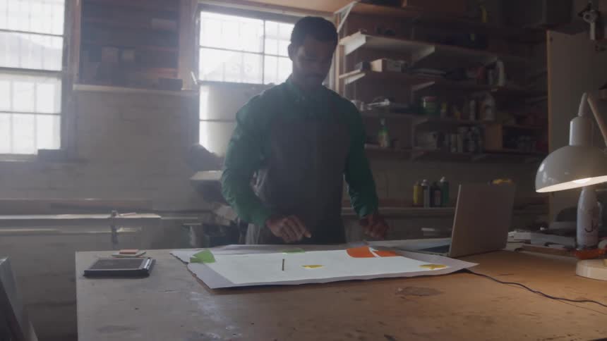 A man in a workshop stands over a lighted work table drawing with a pencil on a large sheet of paper with a laptop and a lamp visible on the table