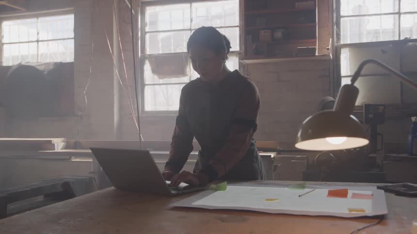 A woman with a bun in her hair and wearing an apron works on a laptop on a dusty workbench in a workshop lit by sunlight