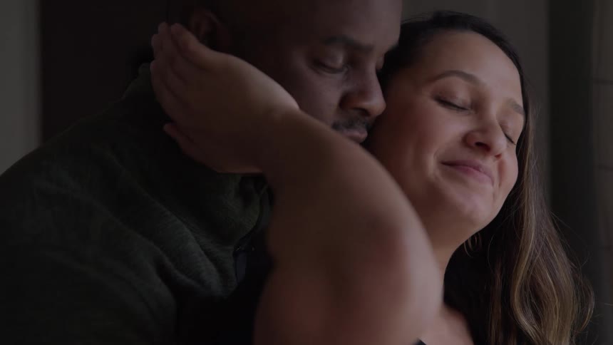 A man and woman stand close together and the man puts his head near the womans neck while she smiles