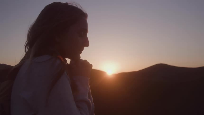 Woman in silhouette stands outdoors at sunset with hands near face against backdrop of hills