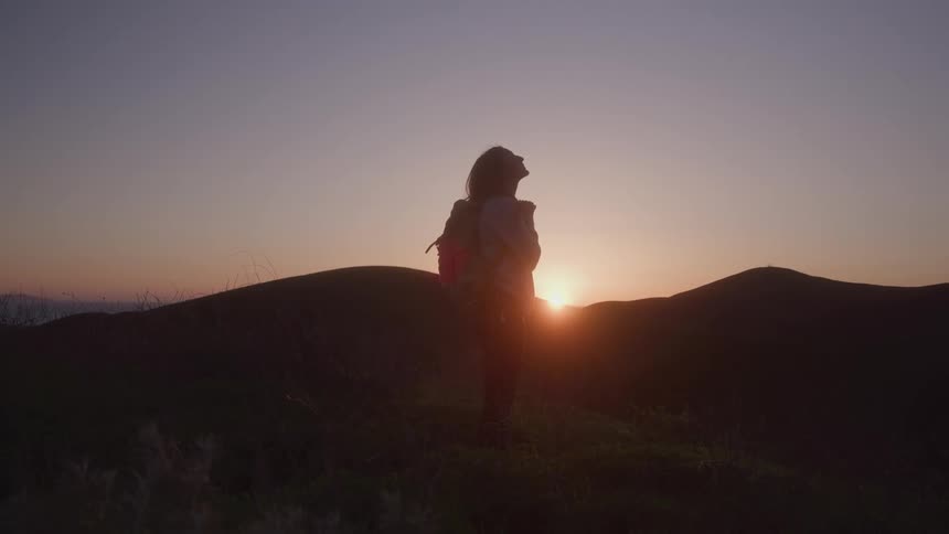A young woman stands on a hill and looks up toward the sky with the sun behind them