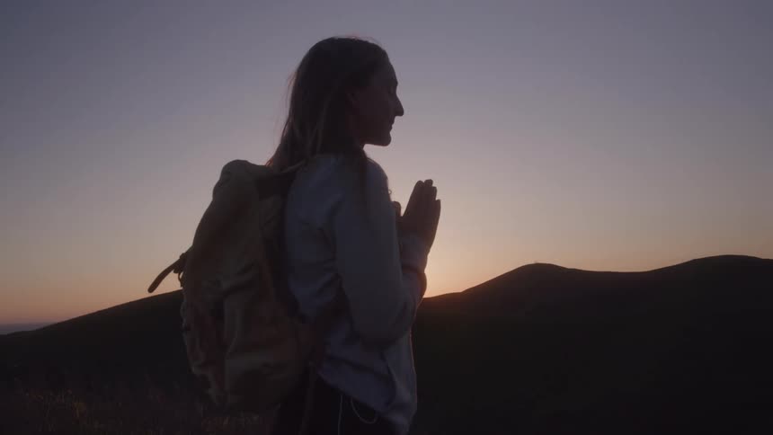 Woman with a backpack makes a prayer gesture facing the sunset