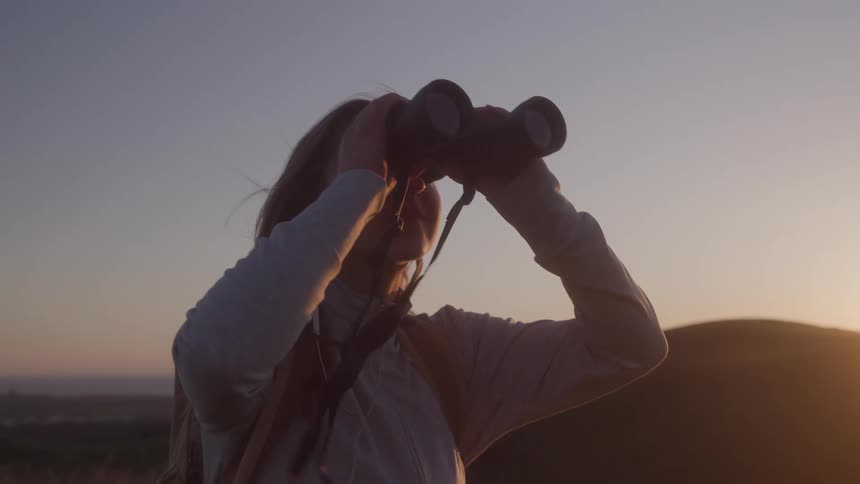 A woman is using binoculars on a hill during a sunset