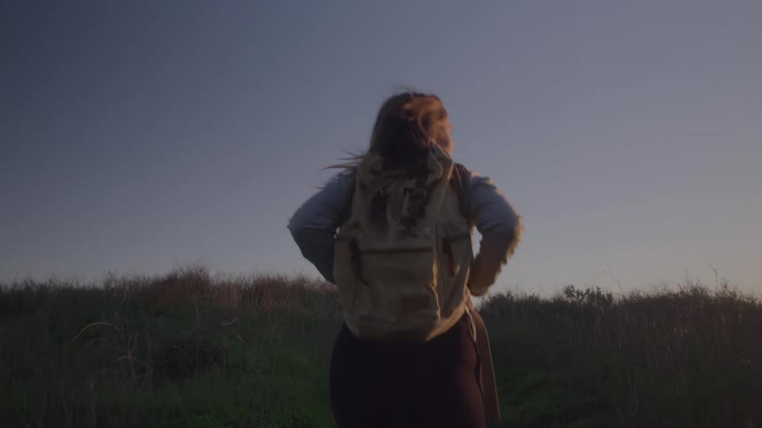 A person stands with a backpack on their back and their hands on their hips looking toward the horizon
