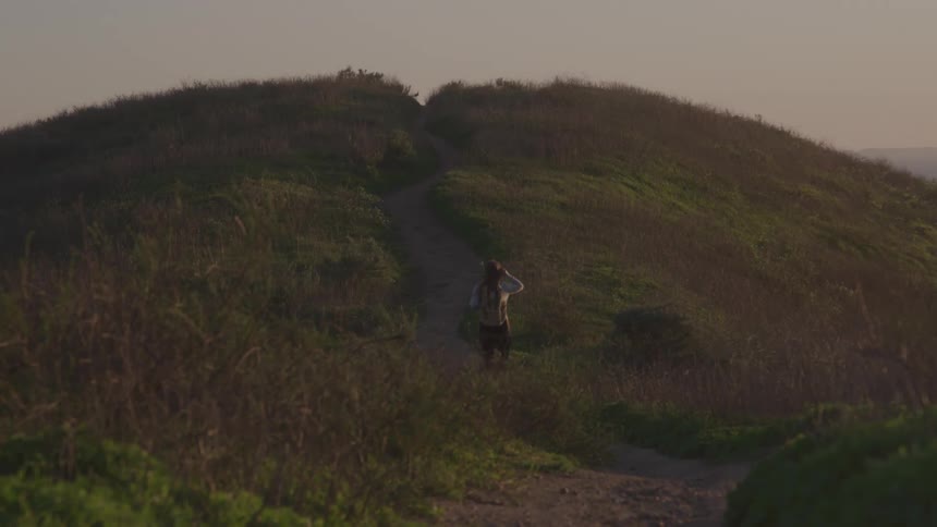A young woman walking through a path on a highland field outdoors at sunset
