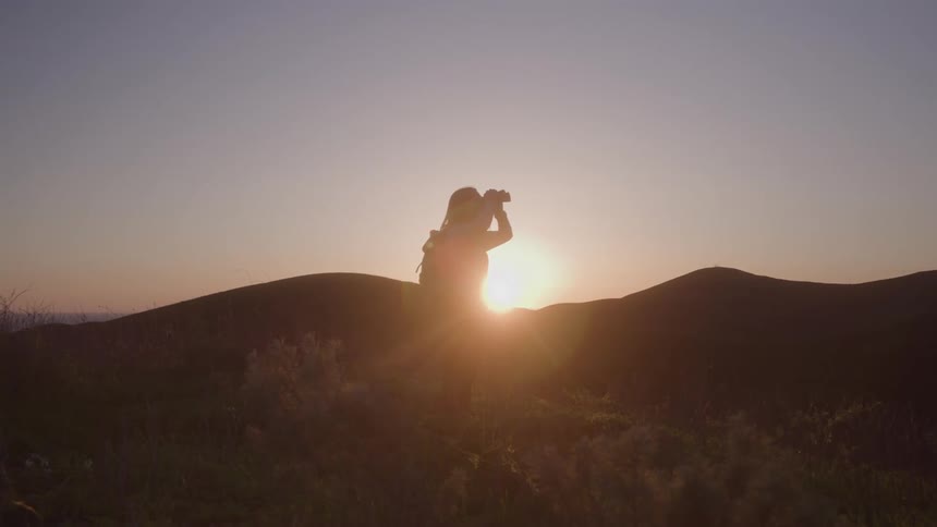 A woman with a backpack stands on a hill with other hills in the distance she lifts binoculars to her eyes and looks to her right