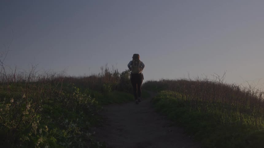 A young woman runs up a hill with a backpack on her back on a clear sky day