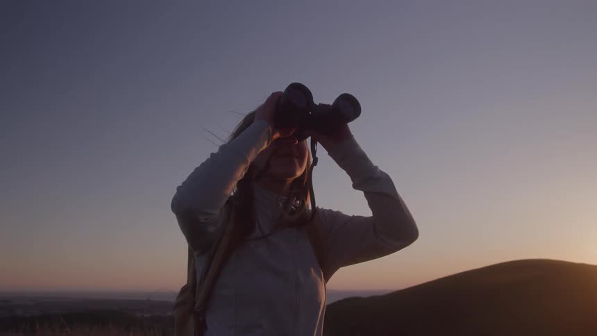 Close up of a young woman viewing her surrounding with a pair of binoculars while standing outdoors at sunset