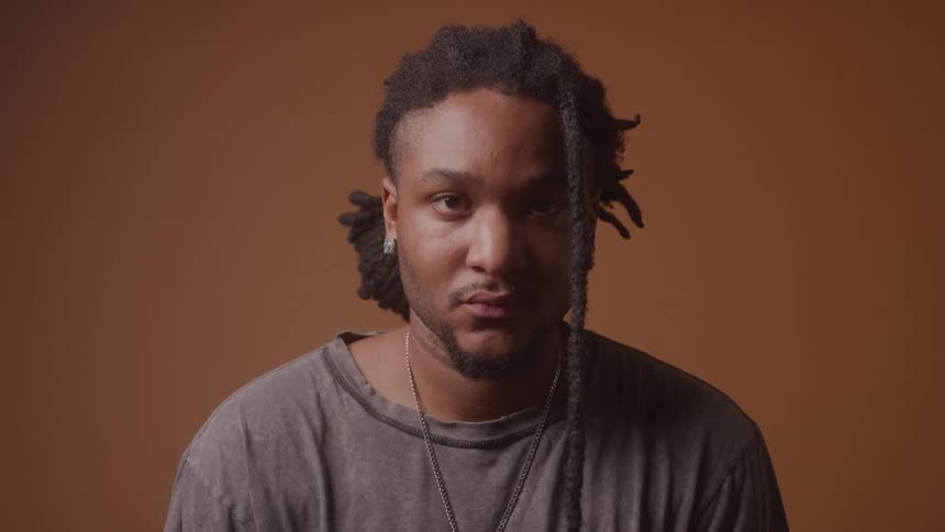 A person with black dreadlocks and a beard is looking down then raises their head to look at the camera they are wearing a grey tshirt and a silver necklace