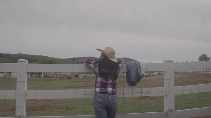 Woman wearing a hat and plaid shirt leaning on a farm fence