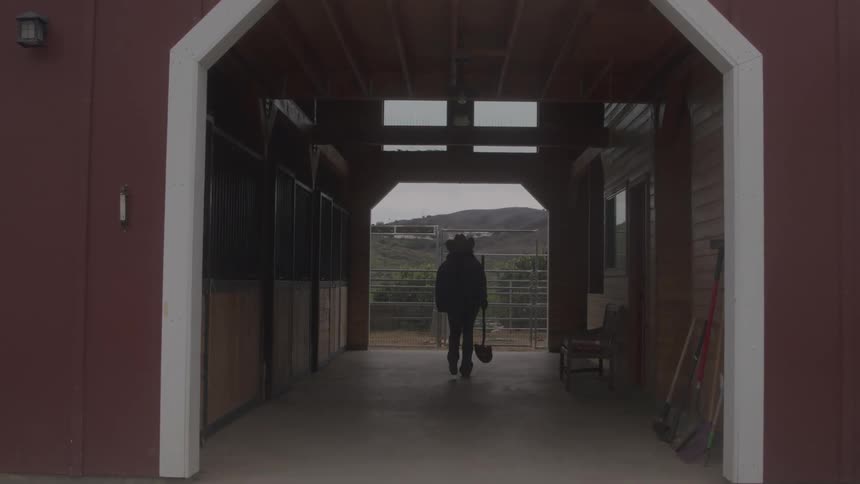 Person walks through stable corridor carrying a shovel between wooden stalls and tools leaning against the wall