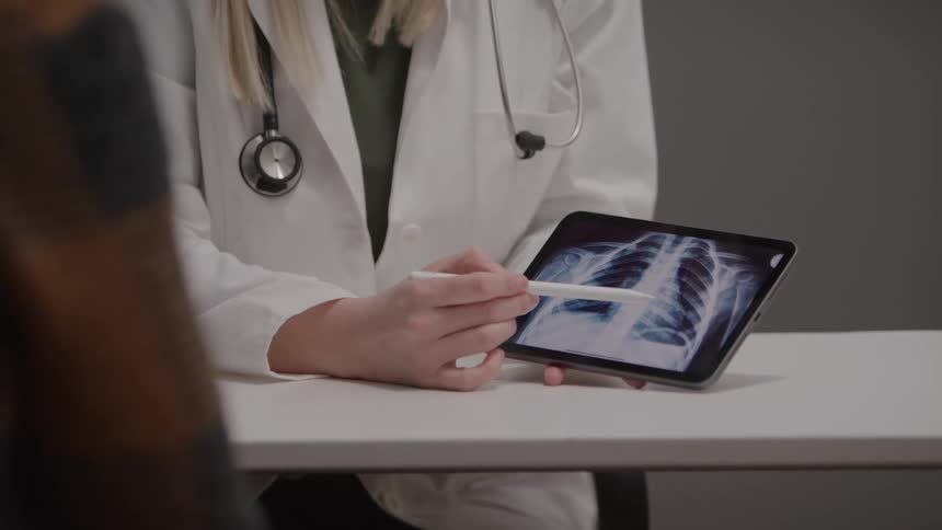 A woman in a lab coat sits at a table and points a stylus at a tablet displaying an xray