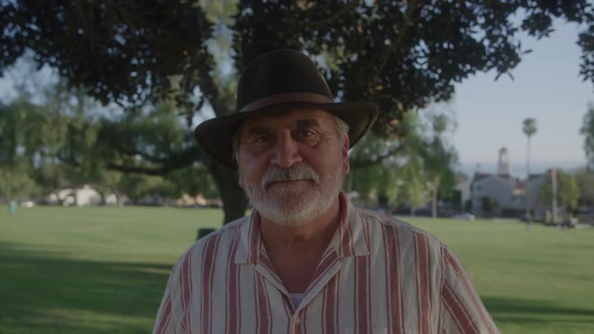Older man in hat and striped shirt standing under a tree at a park