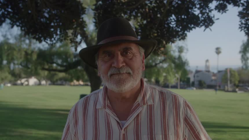 An elderly man with a gray beard and a dark fedora is smiling and looking at the camera wearing a red and white striped shirt in a sunny park setting