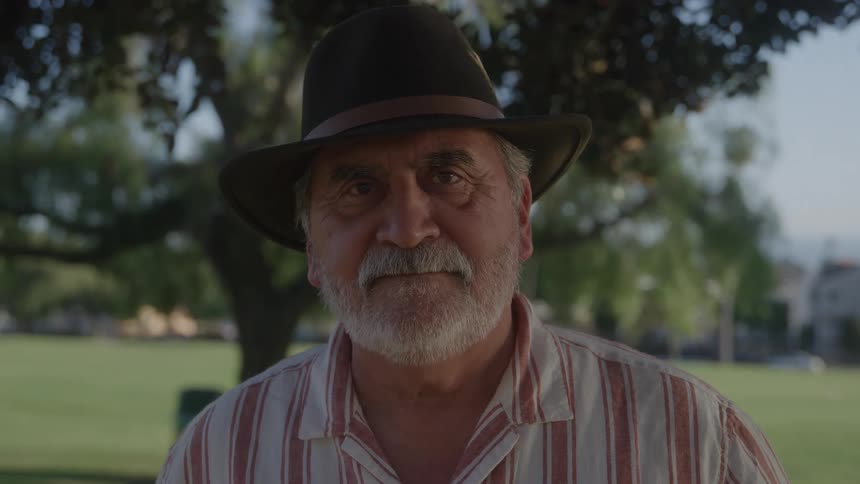 Close up of older man in a hat and striped shirt near a tree at a park